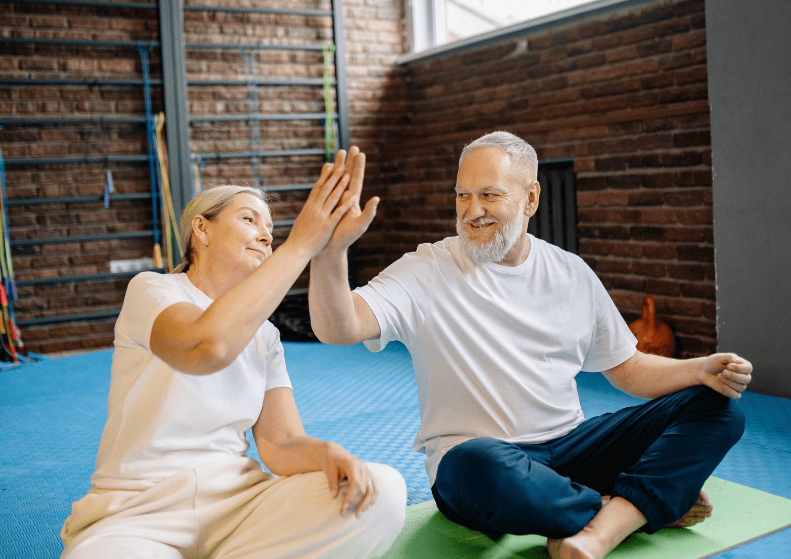 Older couple sitting cross-legged on exercise mats and high-fiving during a workout in a brick-walled gym.