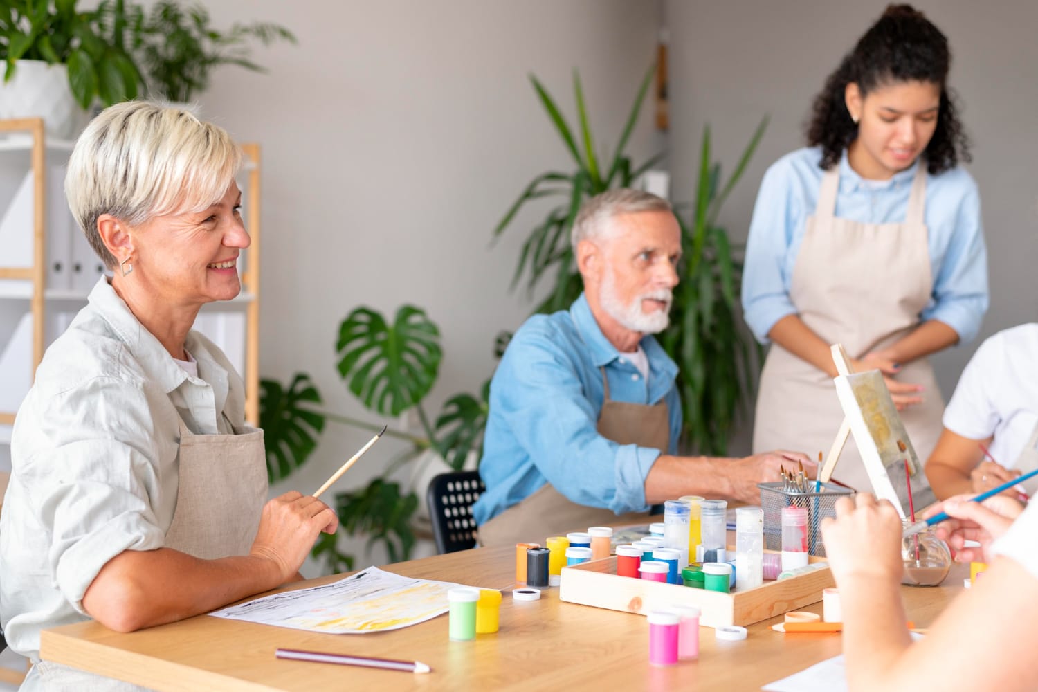 Older adults and a caregiver paint together at a bright art table, sharing brushes and colorful paints in a cheerful, welcoming workshop.