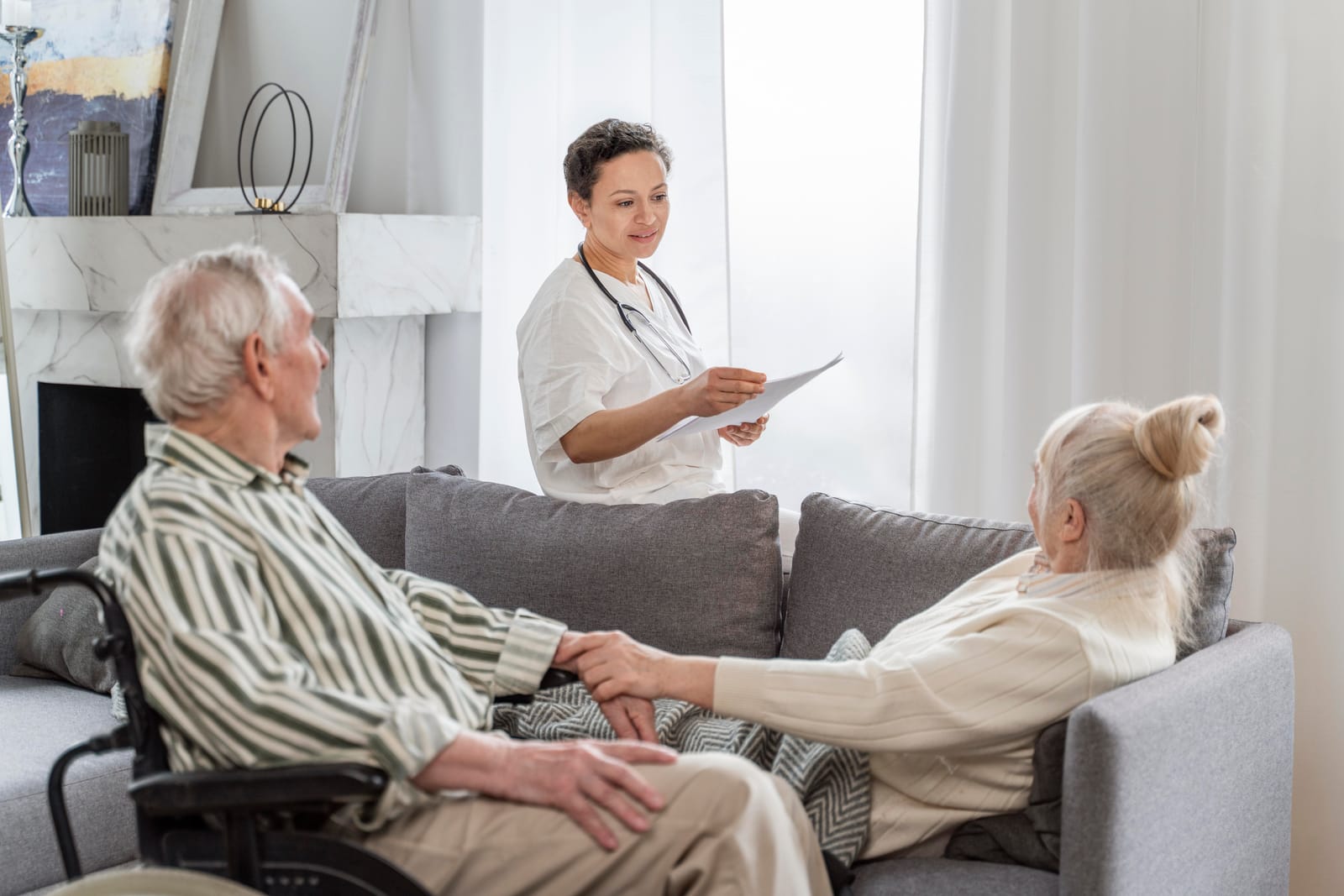 A female healthcare worker in a white uniform with a stethoscope talks to an elderly couple sitting on a gray sofa in a bright, modern living room.