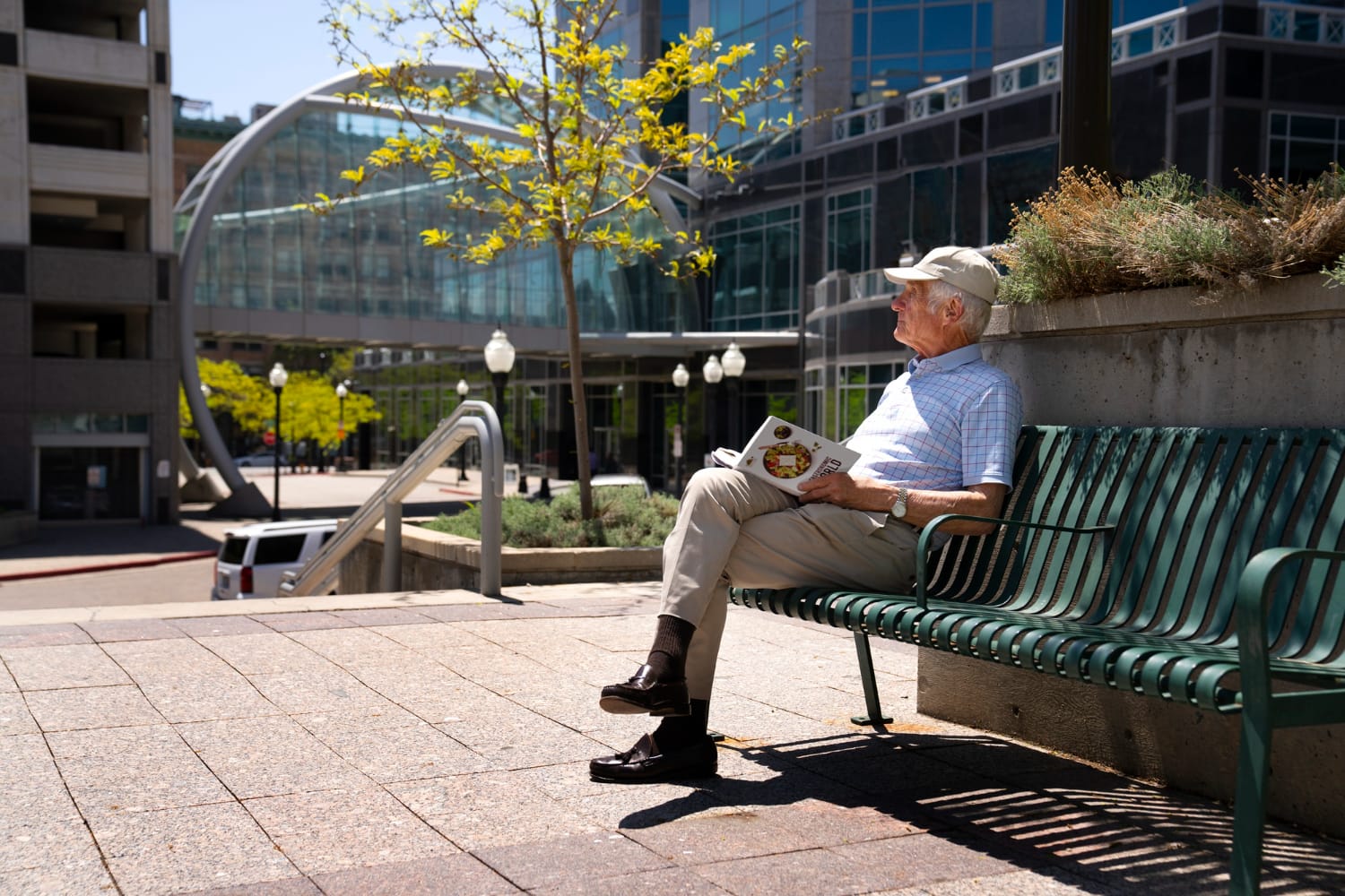 A man sitting on a wooden bench in a park, wearing a blue shirt and jeans, looking at his phone.