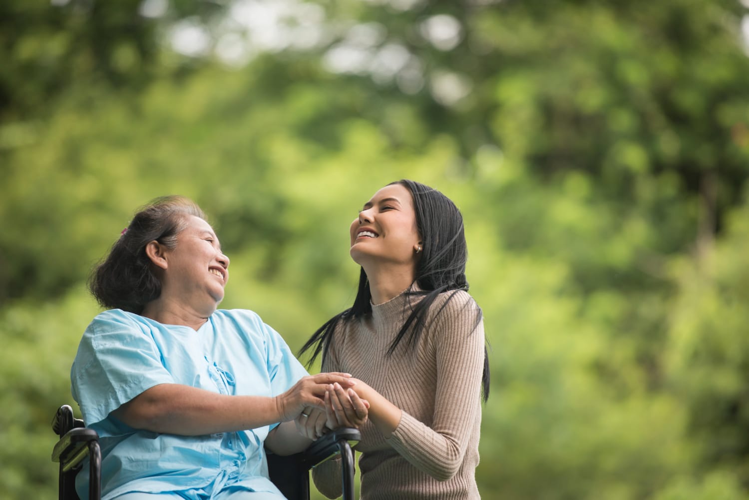 An older woman in a wheelchair smiles and holds hands with a younger woman outdoors.
