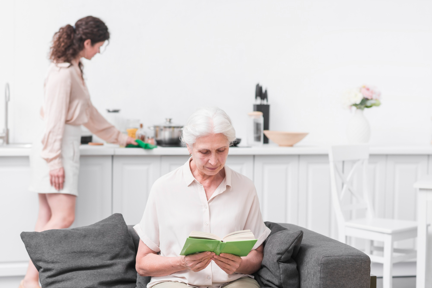 An elderly woman with gray hair sits on a gray sofa reading a green book, while a younger woman in the background cleans a white kitchen counter.