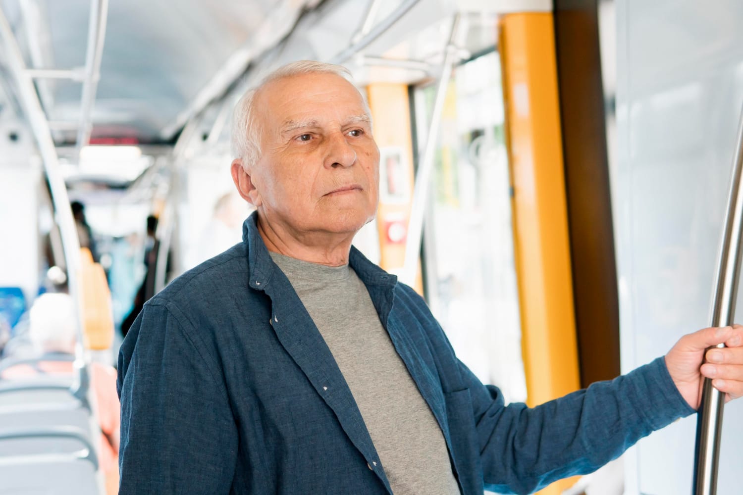 An older man stands inside a bus, holding onto a handrail for support.