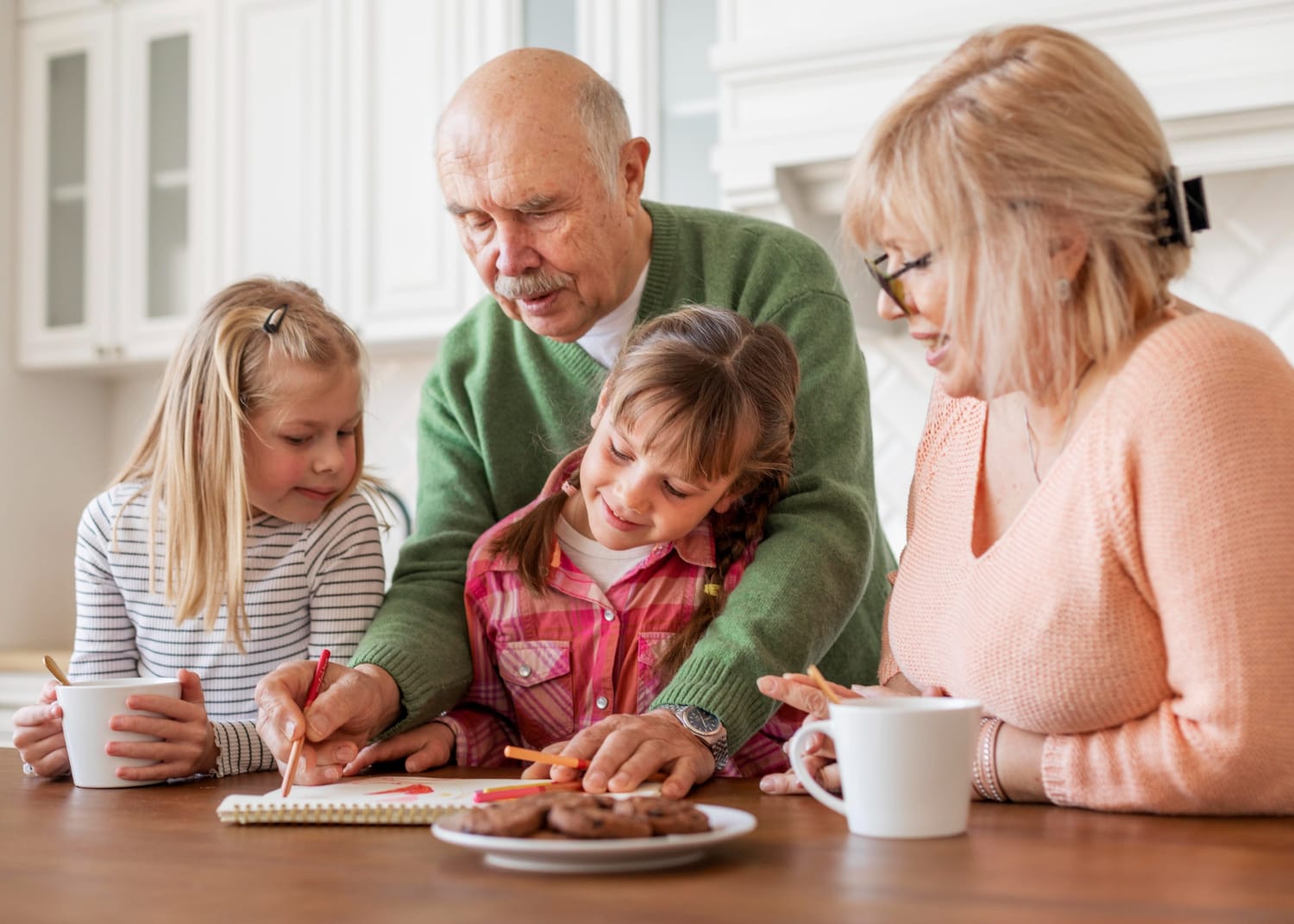 An elderly man and woman assist two young girls with drawing on a notepad at a wooden table, with cookies and cups nearby.