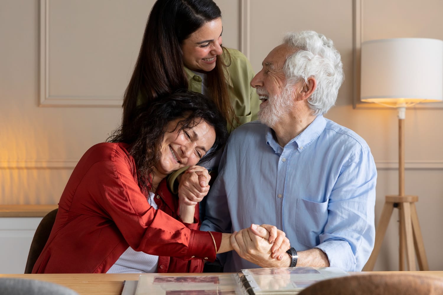 A smiling young woman stands behind an older couple, who are laughing and holding hands while looking at a photo album on a wooden table indoors.