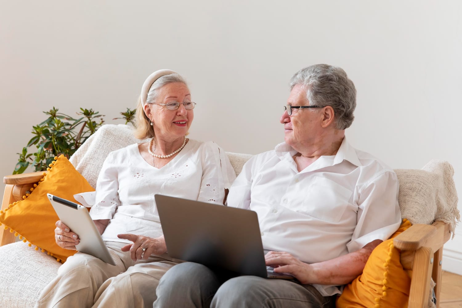 An elderly couple sits on a couch, with the woman holding a tablet and the man using a laptop, both looking at each other in a bright living room.