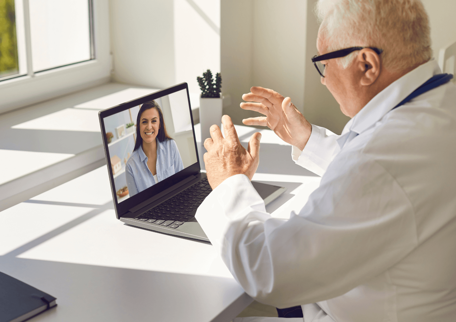 Doctor in a white coat gesturing during a video call with a patient on a laptop screen.