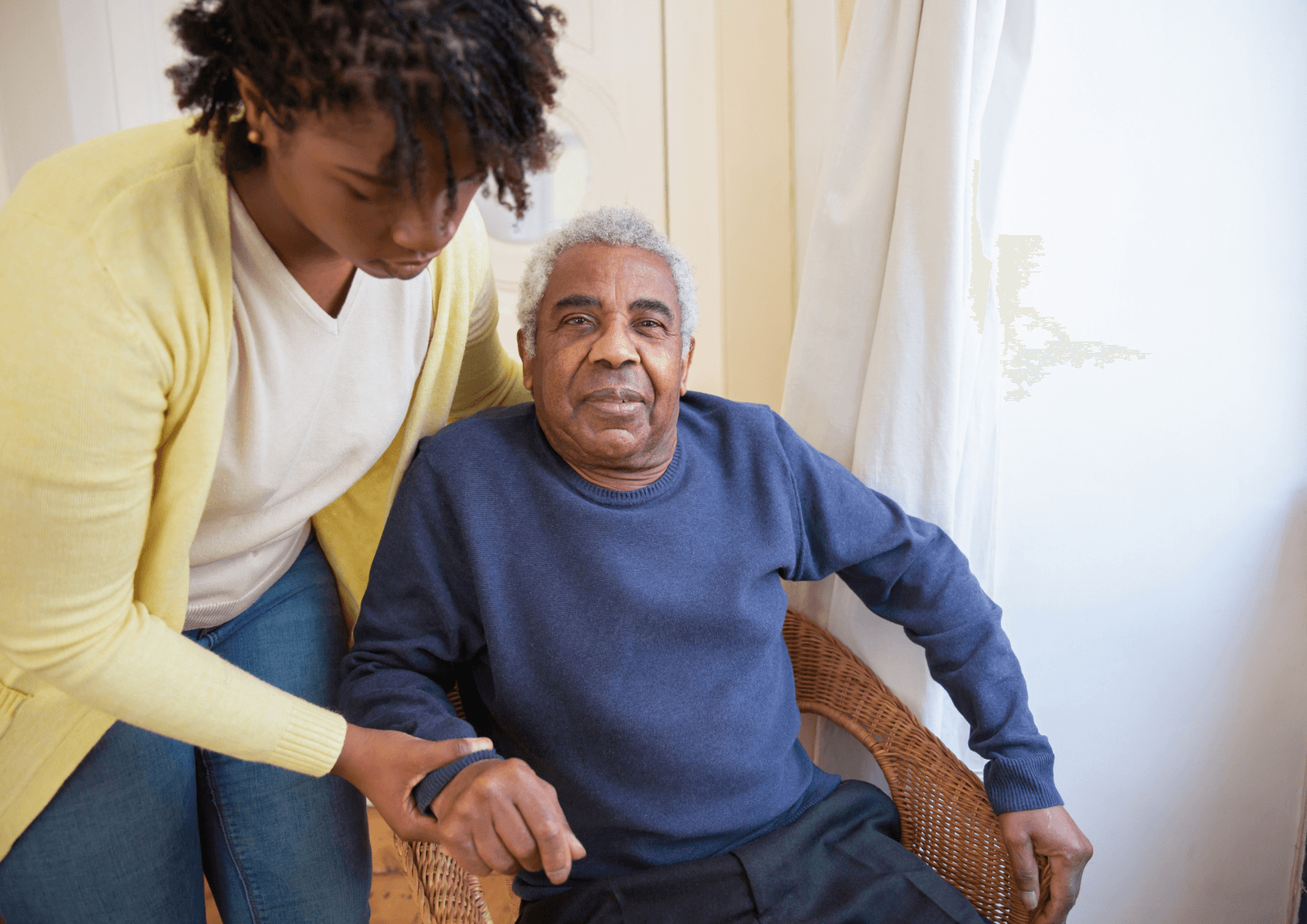 Younger person leaning in to help an older man seated in a wicker chair beside a window.