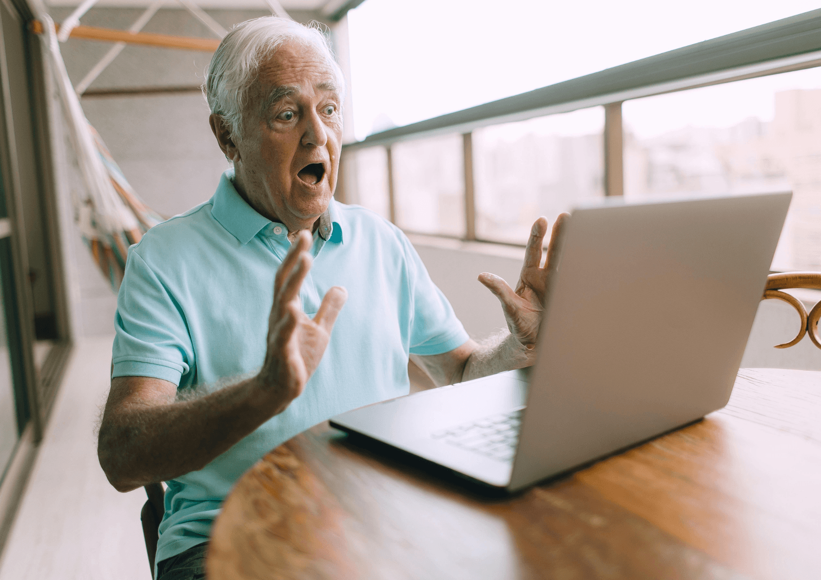 Older man looking shocked with hands raised while seated at a table in front of an open laptop.