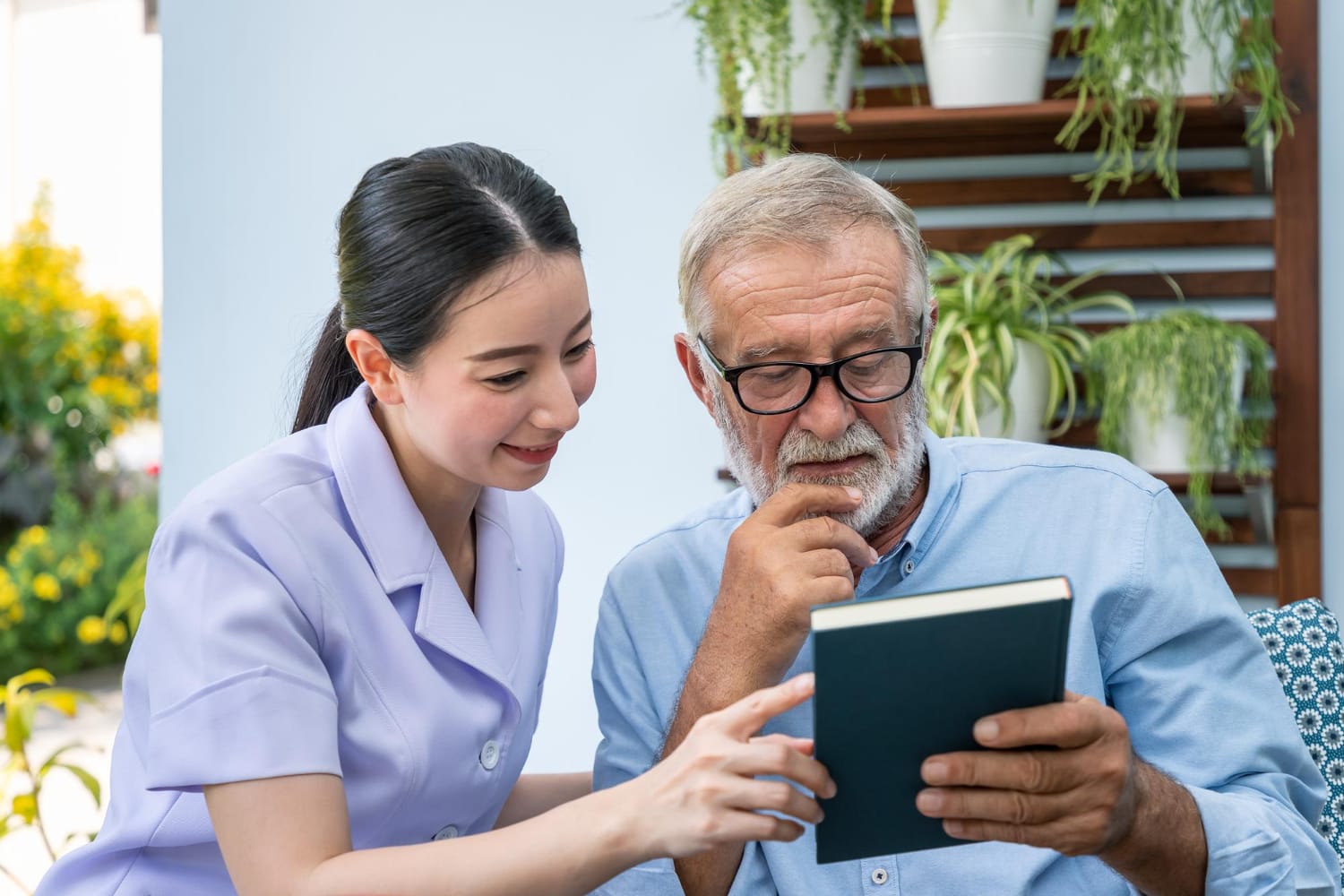 A young female caregiver in a light purple uniform points at a small book held by an elderly man with a grey beard and glasses in a garden setting.