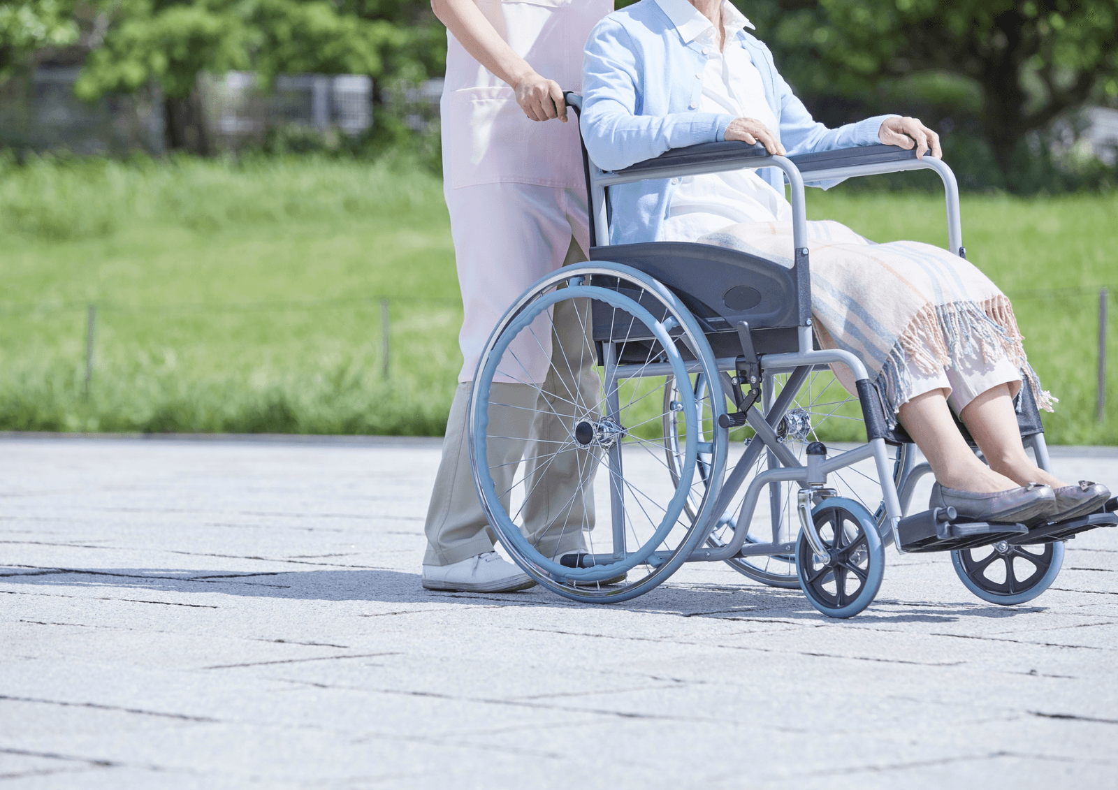 Caregiver in scrubs pushing an older adult in a wheelchair along a paved path outdoors.