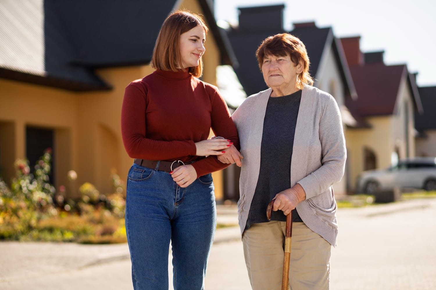 A young woman in a red turtleneck and jeans walks with an elderly woman in a gray cardigan.