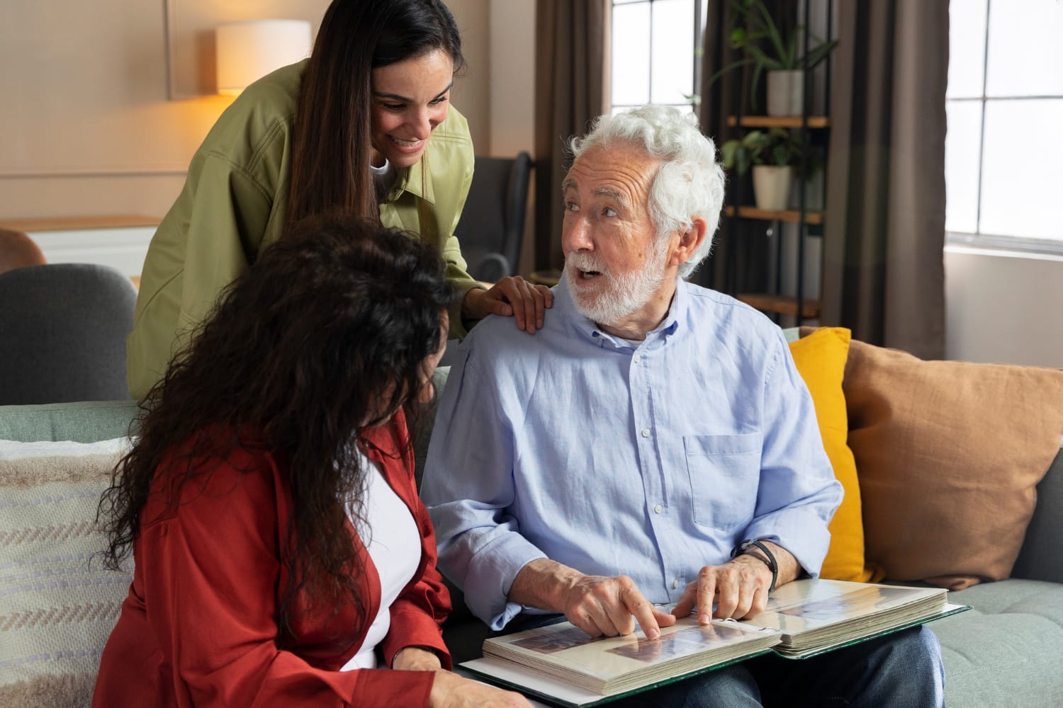 A young woman in a green shirt leans over a sofa, smiling at an elderly man and a woman in red who are looking at a photo album together indoors.