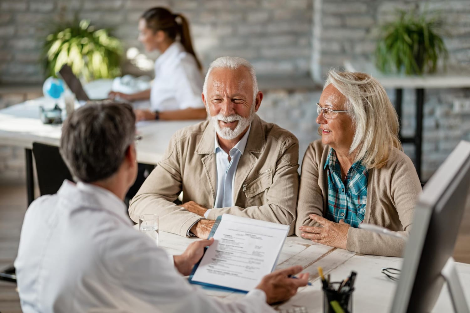 An older couple smiles as a professional reviews paperwork with them across an office desk, creating a warm, reassuring consultation.