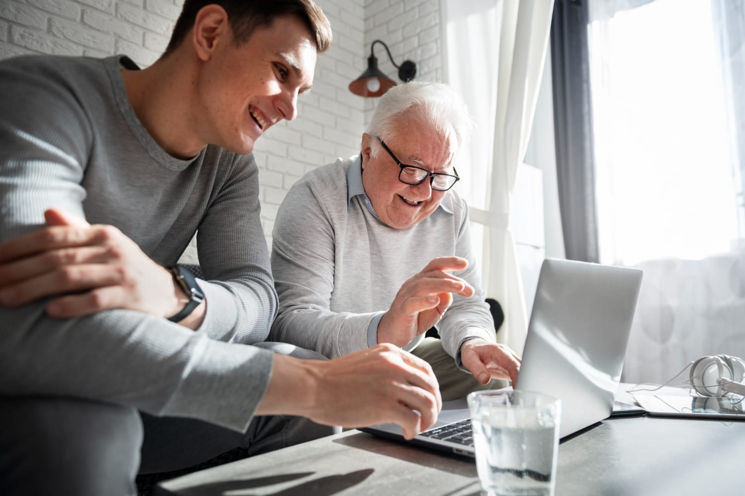 A smiling young man types on a laptop beside an older man gesturing toward the screen in a bright office, sharing a warm, collaborative moment.