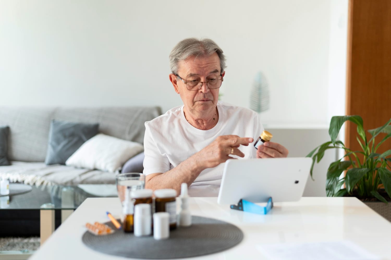 An elderly man at a table, using a tablet with a bottle of pills nearby.