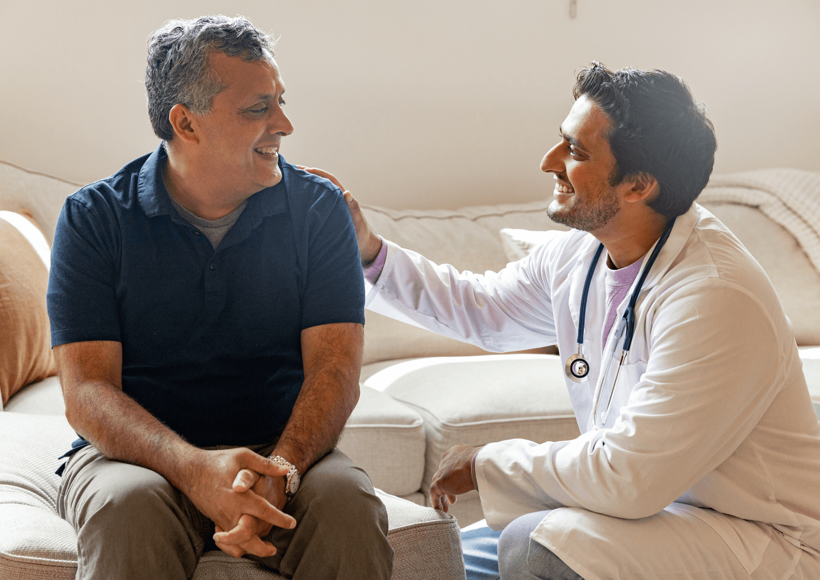 Doctor in a white coat with a stethoscope sitting on a couch, resting a hand on a patient's shoulder while they talk.