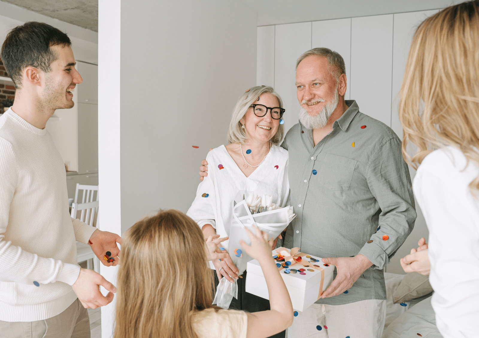 Older couple holding flowers and a gift box surrounded by family members tossing confetti indoors.