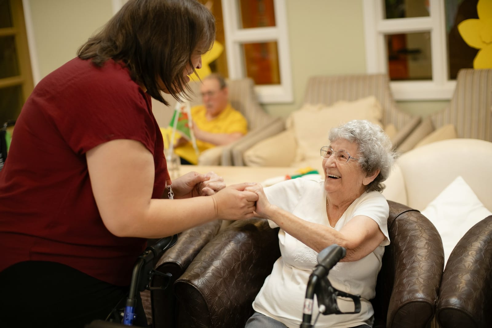 A caregiver in a maroon shirt holds hands with an elderly woman in a white shirt, who sits in a brown armchair with a walker in a bright room.