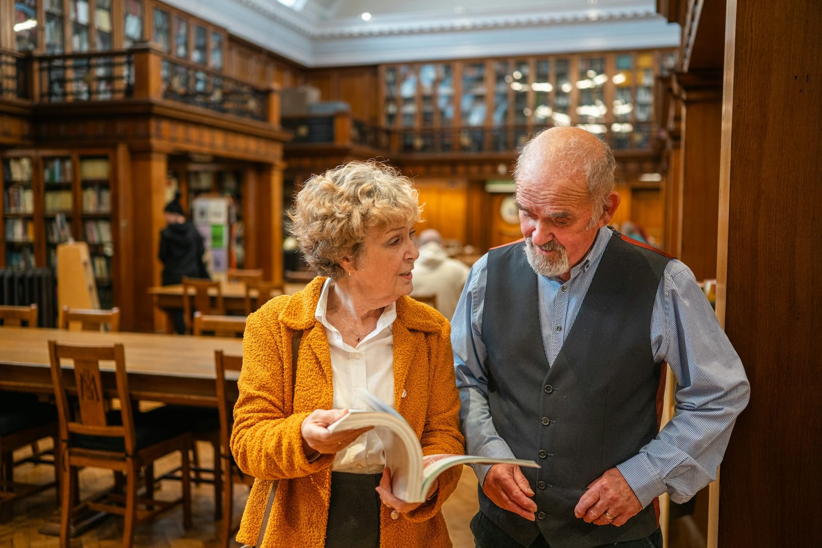 An older woman in a yellow jacket and a man in a vest stand in a wood-paneled library, looking at an open book together near wooden tables.