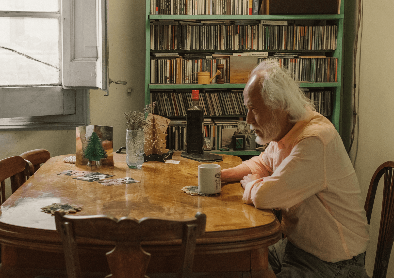 Older man with white hair sitting alone at a wooden table with a mug, in front of a shelf full of CDs.