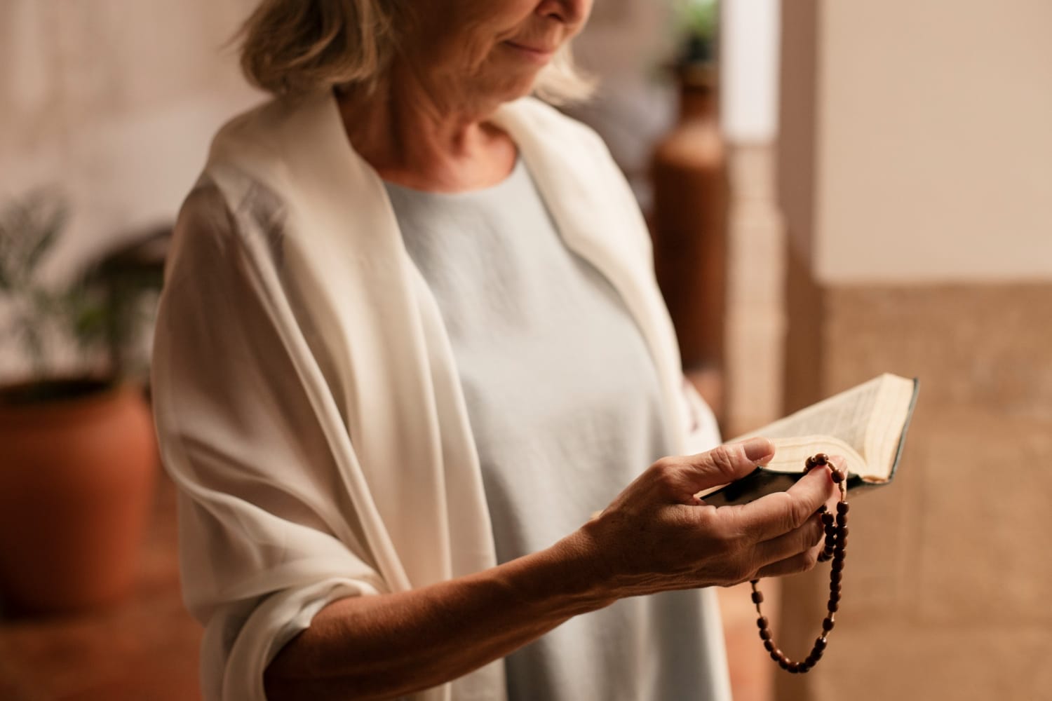 A woman holding a book in one hand and a rosary in the other, wearing a light-colored shawl, in a softly lit indoor setting.