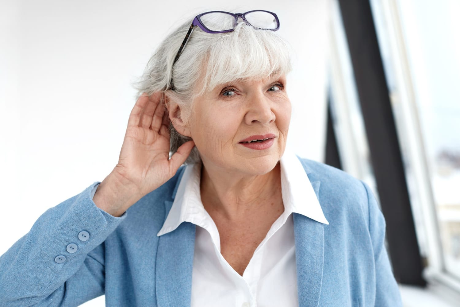 An older woman in a blue jacket with glasses holds her hand to her ear, listening intently.