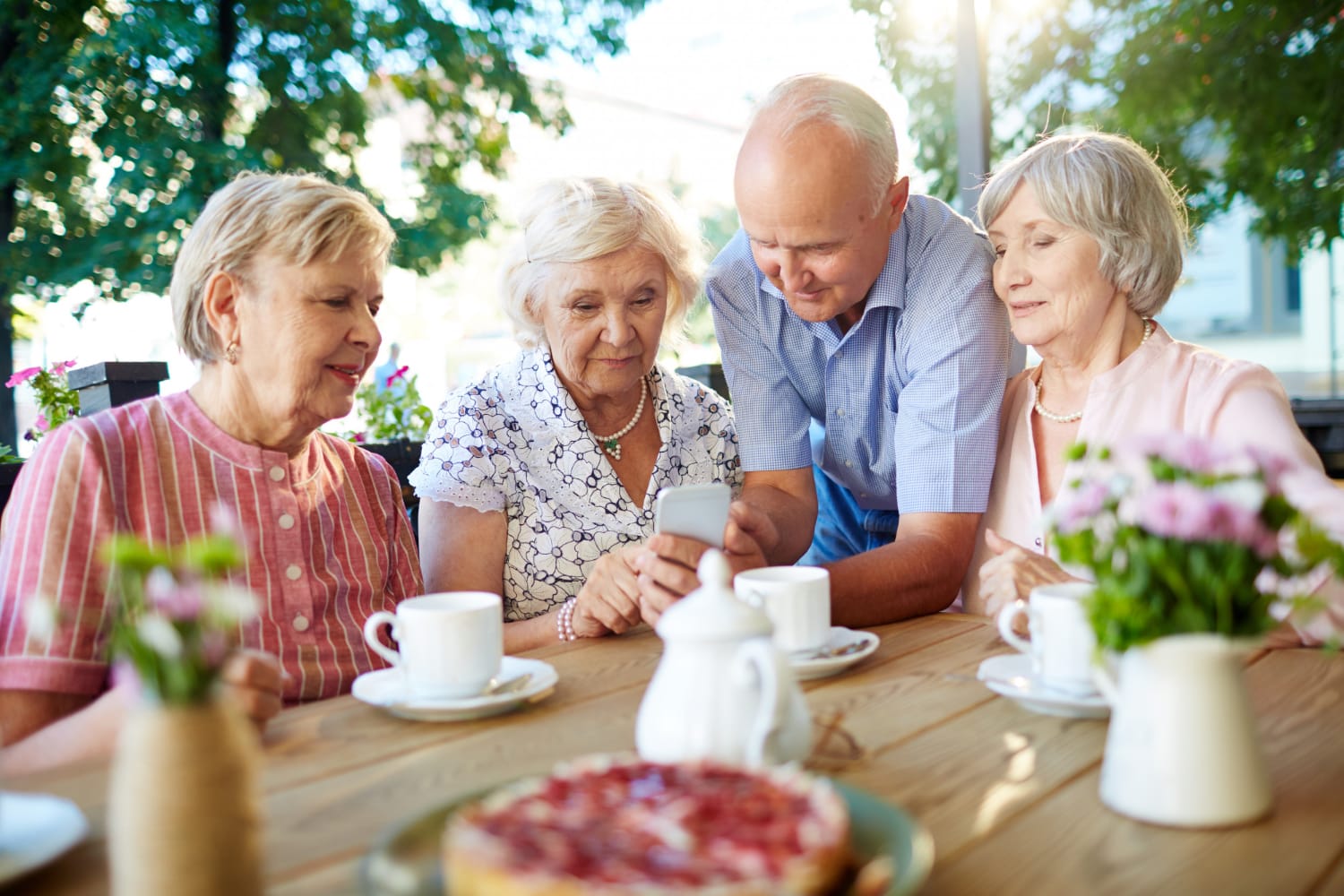 Four older adults gathered around a table, looking at a smartphone, with coffee cups and a cake.