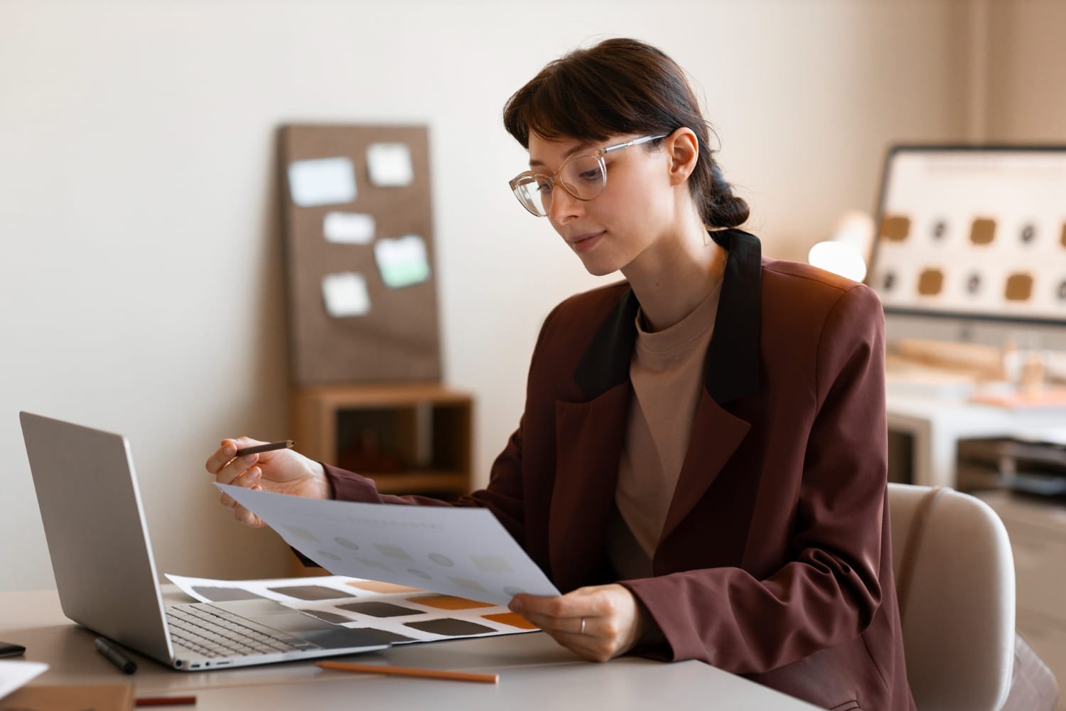 A woman with glasses wearing a brown blazer sits at a desk, reviewing a document with color swatches while looking at her open silver laptop.