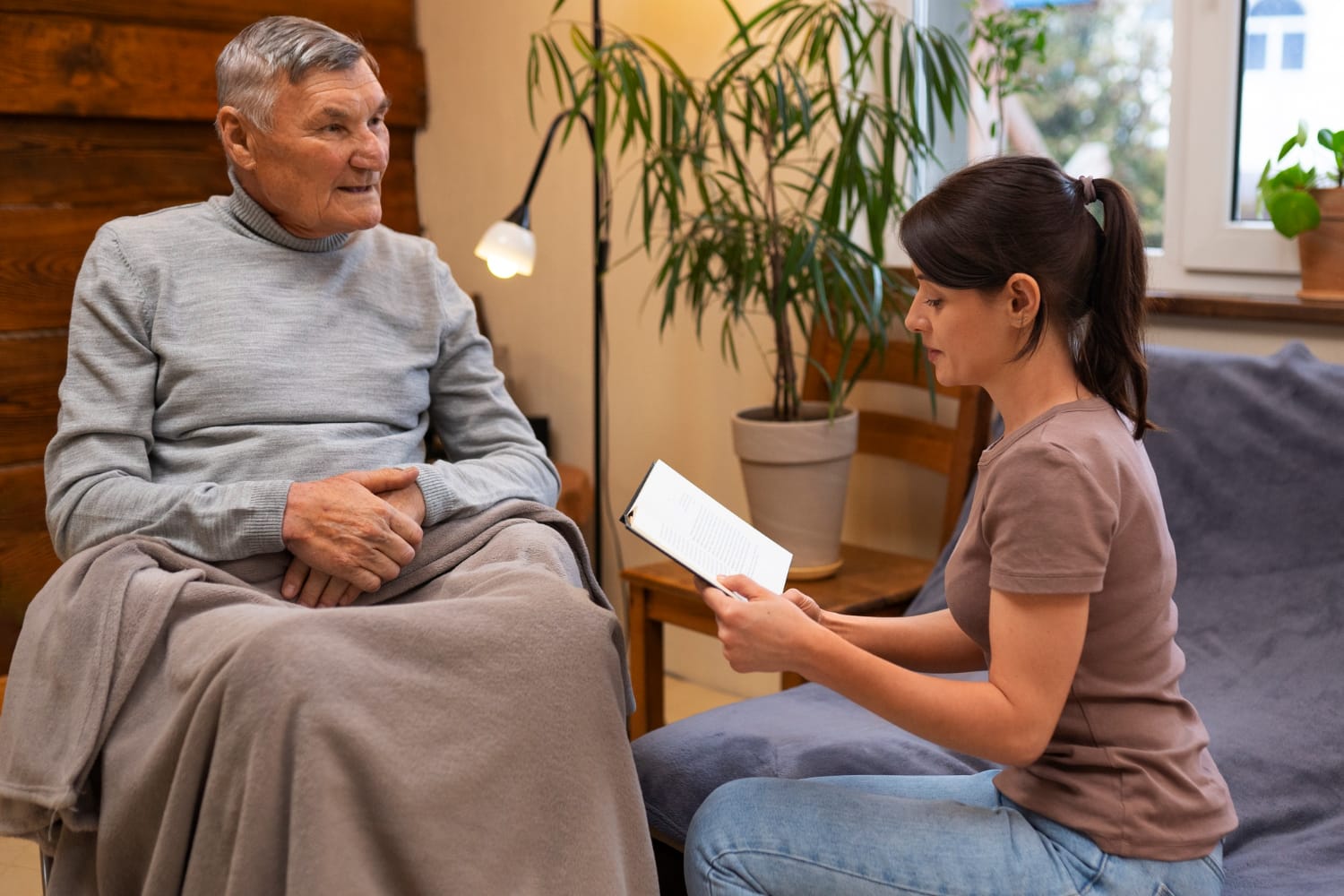 A young woman sits on a sofa, reading a book aloud to an elderly man who is resting under a gray blanket.