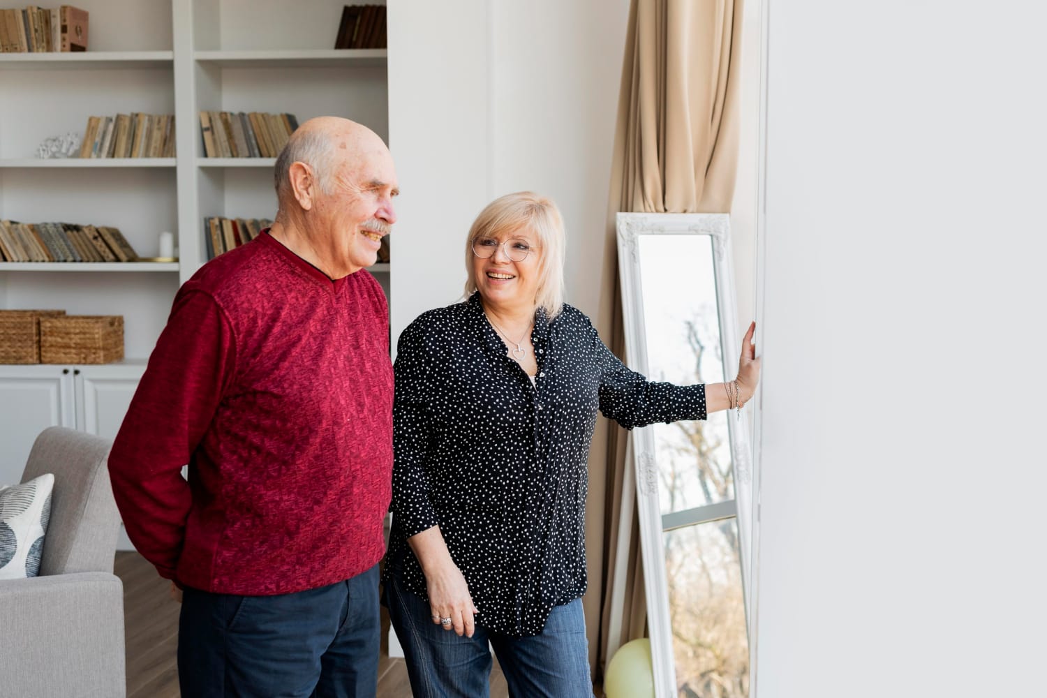 A senior couple smiles at their reflection in a mirror, standing close together.