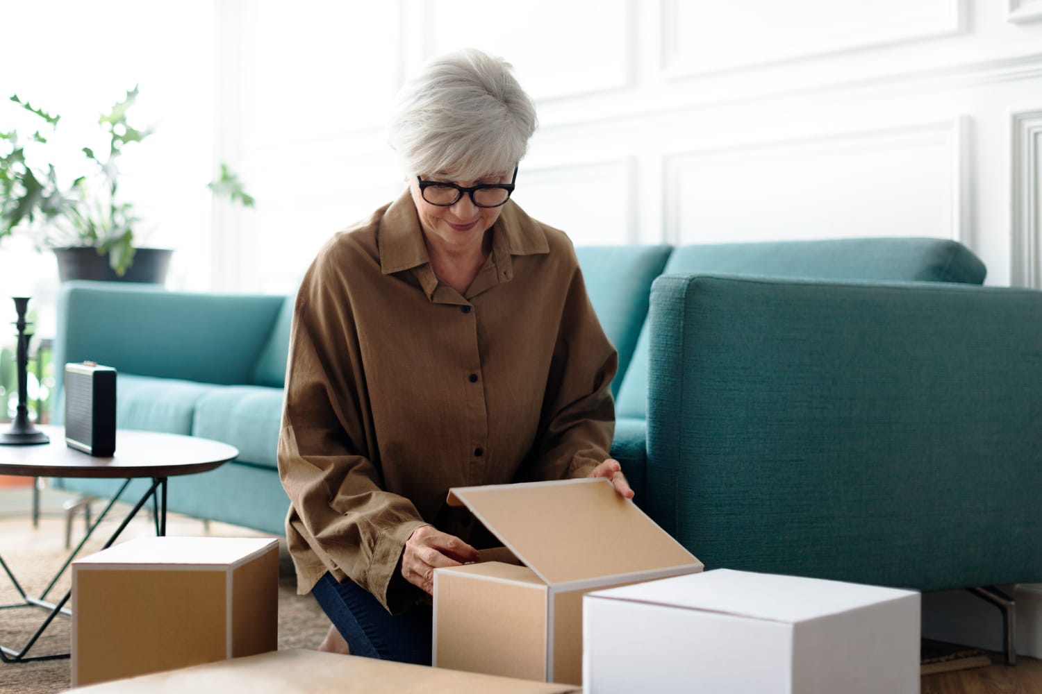 A woman sits on a couch surrounded by cardboard boxes, looking thoughtful.
