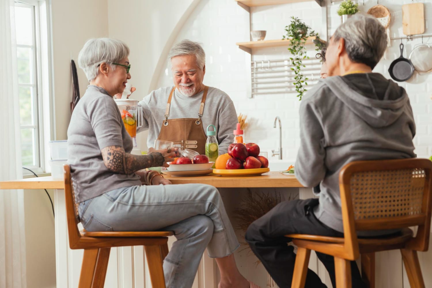 Three elderly people sit at a kitchen island, with one man wearing an apron preparing a smoothie while others watch.