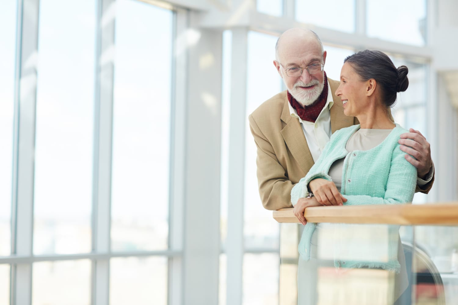 An elderly man with a white beard and glasses wearing a tan blazer and a woman in a light green jacket stand together by a bright glass railing.