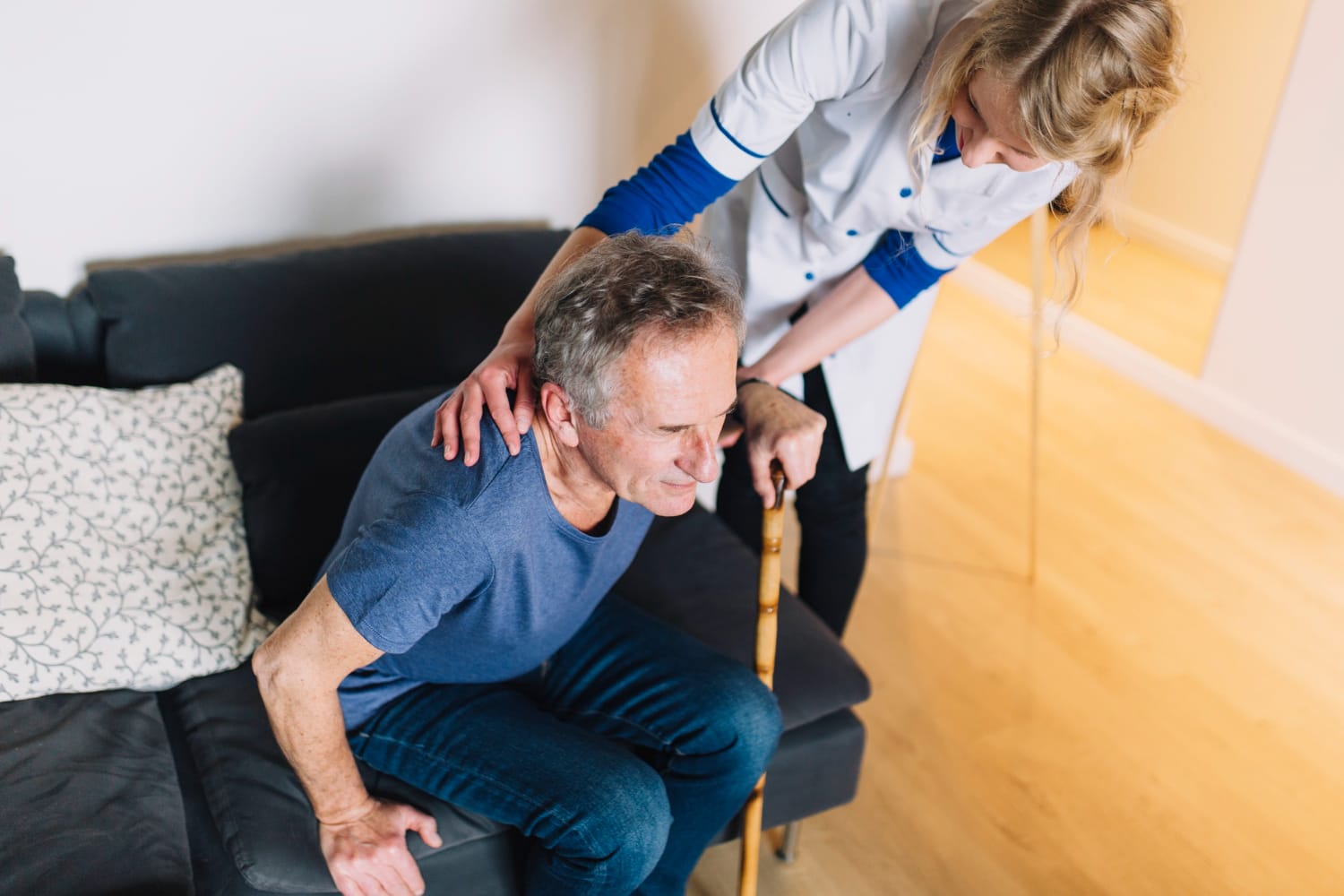 A female caregiver in a white uniform assists an elderly man with a wooden cane as he stands up from a dark gray sofa in a brightly lit living room.