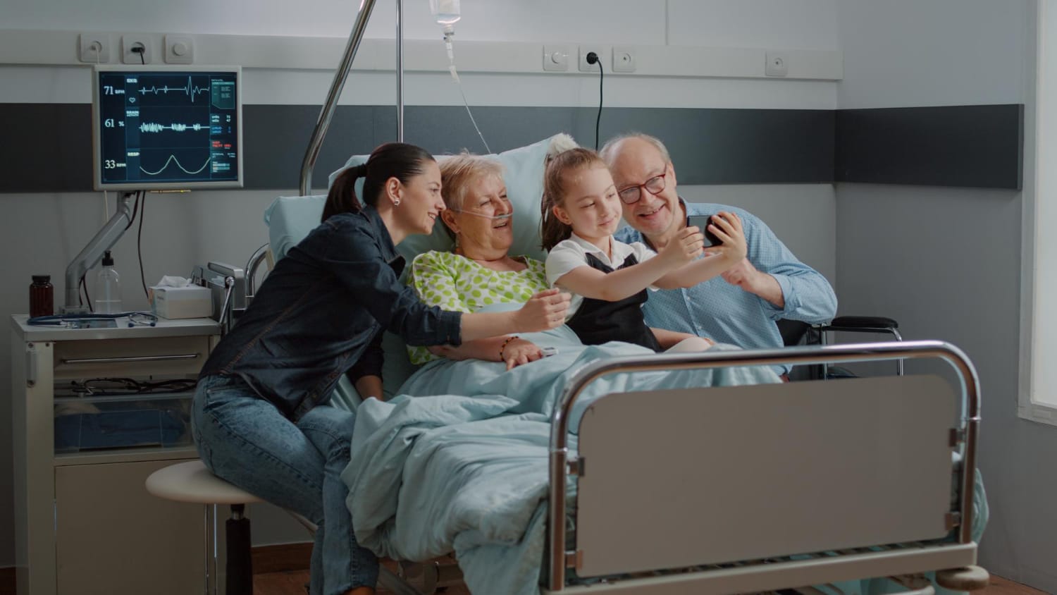 Kid taking selfie on smartphone with ill patient and family at visit in hospital ward bed. Child using mobile phone for pictures and visiting aged woman with old man and mother at clinic.