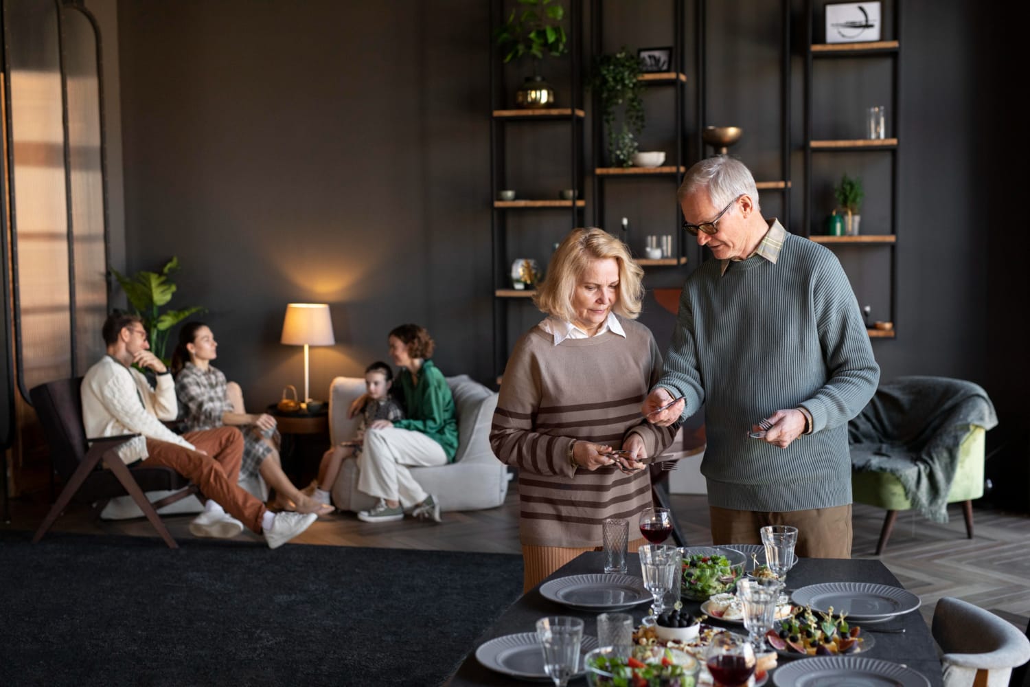 An older couple shares wine beside a dinner table in a warmly lit living room, while friends talk in the background; the mood is relaxed and social.