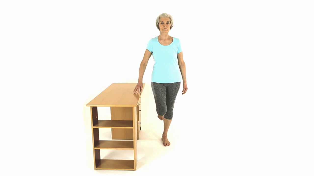 Older adult balancing on one leg while resting a hand on a table for support.