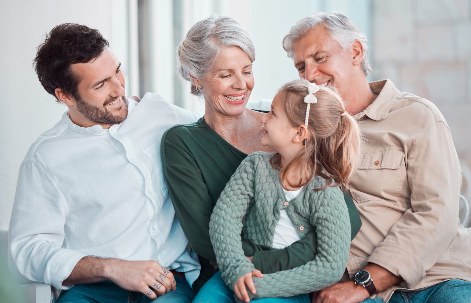 Three generations of a family smiling together on a couch — a young father, grandparents, and a little girl
