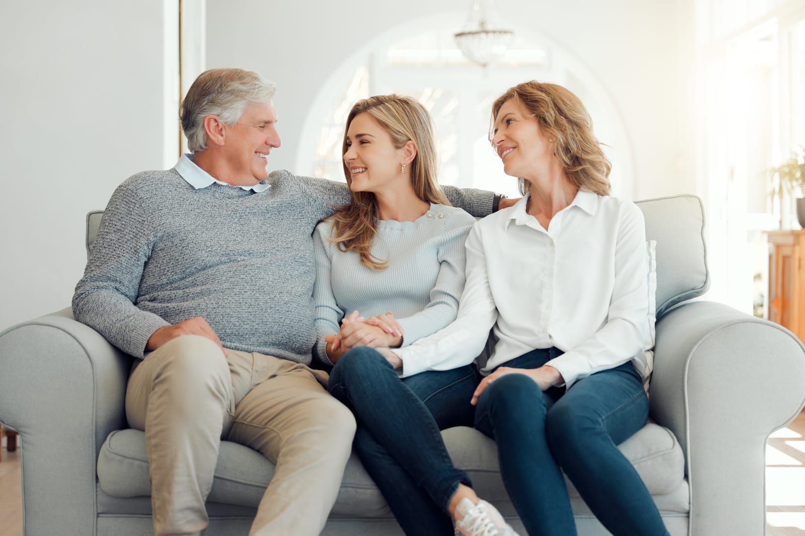 An adult daughter sitting on a couch holding hands and laughing with her two aging parents